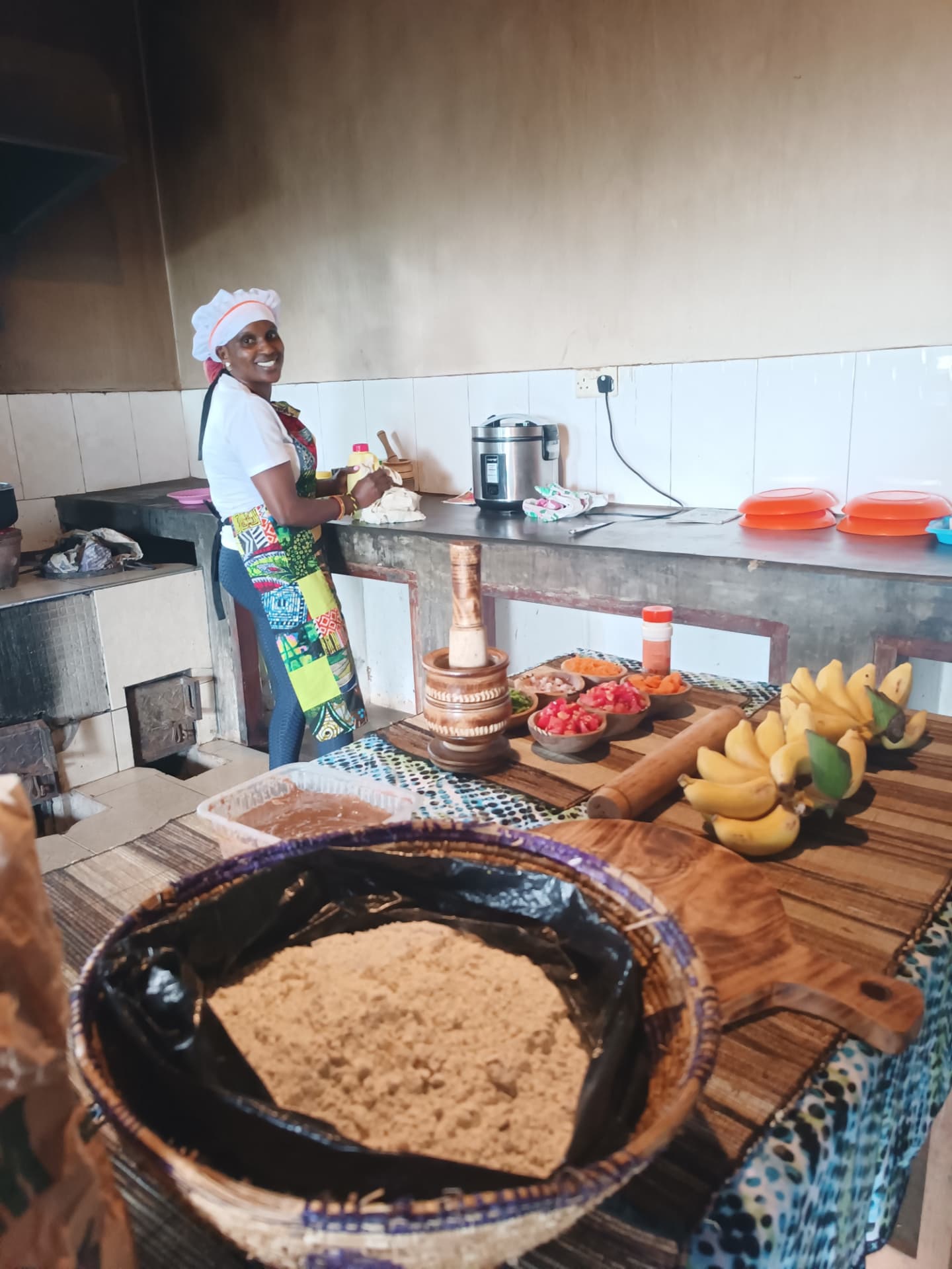 Aine smiling in her kitchen while preparing dough, with bananas, tomatoes and spices laid out on the table